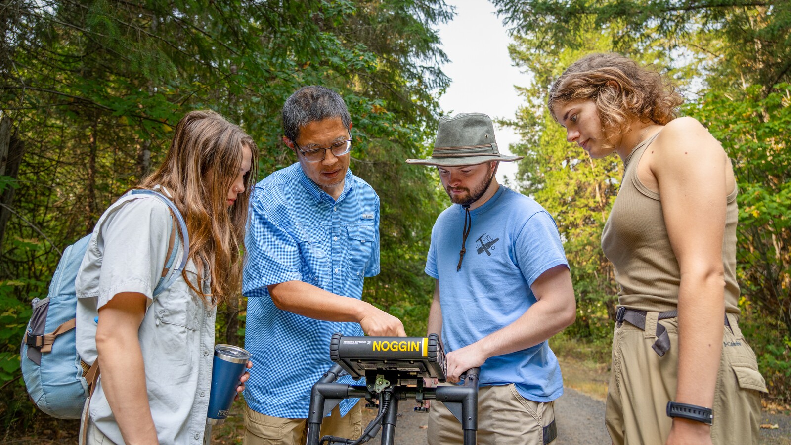 Four people examine field research equipment on a forest road.