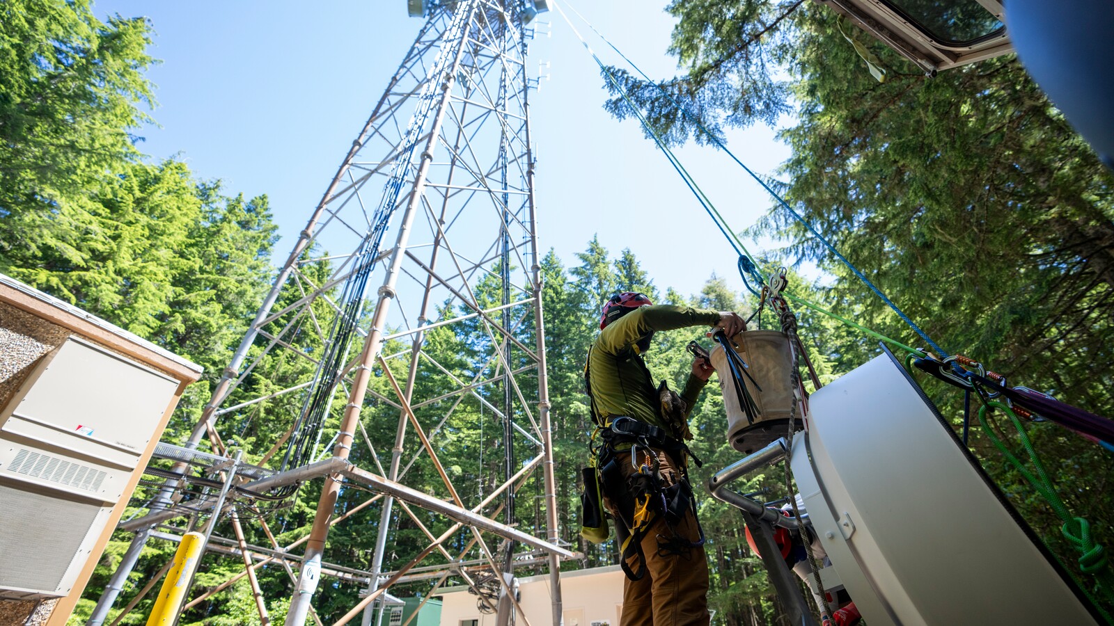 A technician installs wildfire camera equipment beside a forest communications tower.