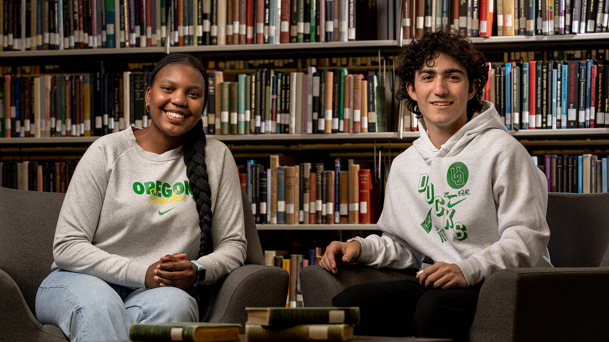 Two students sit in armchairs in front of library stacks.