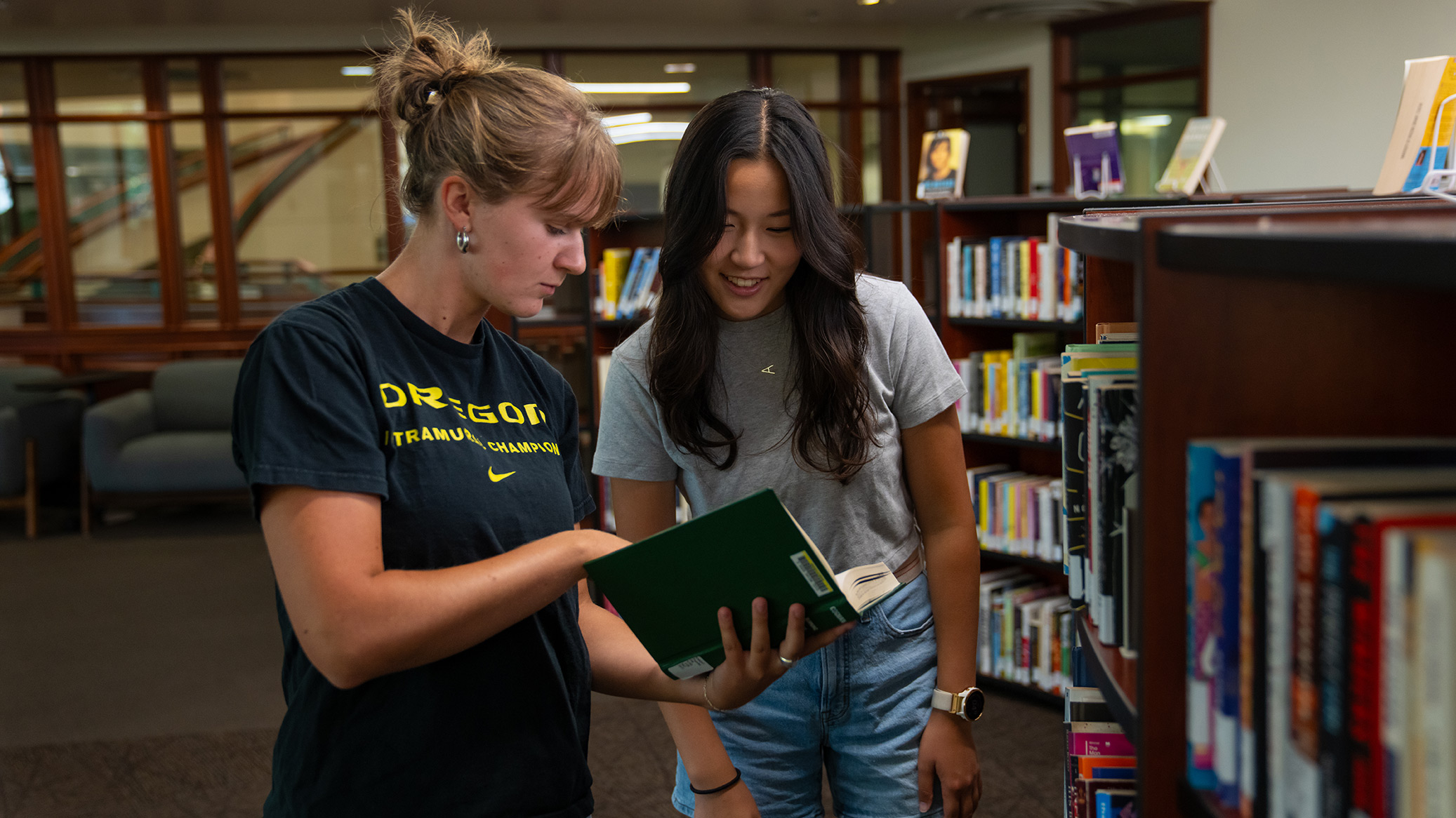 Two students look through a book among library shelves.