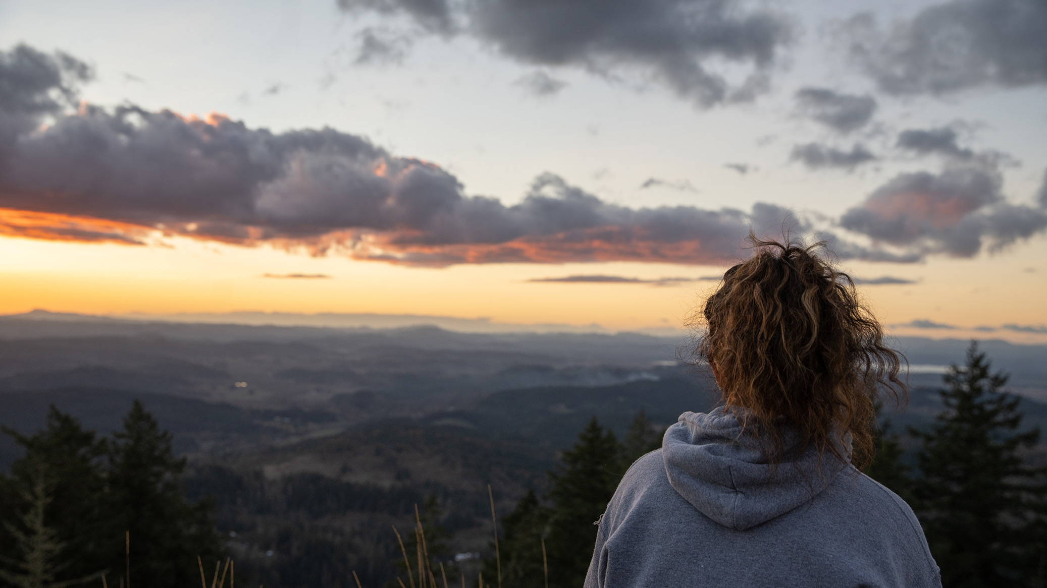 A person in a gray hoodie looks out over a valley at sunset.