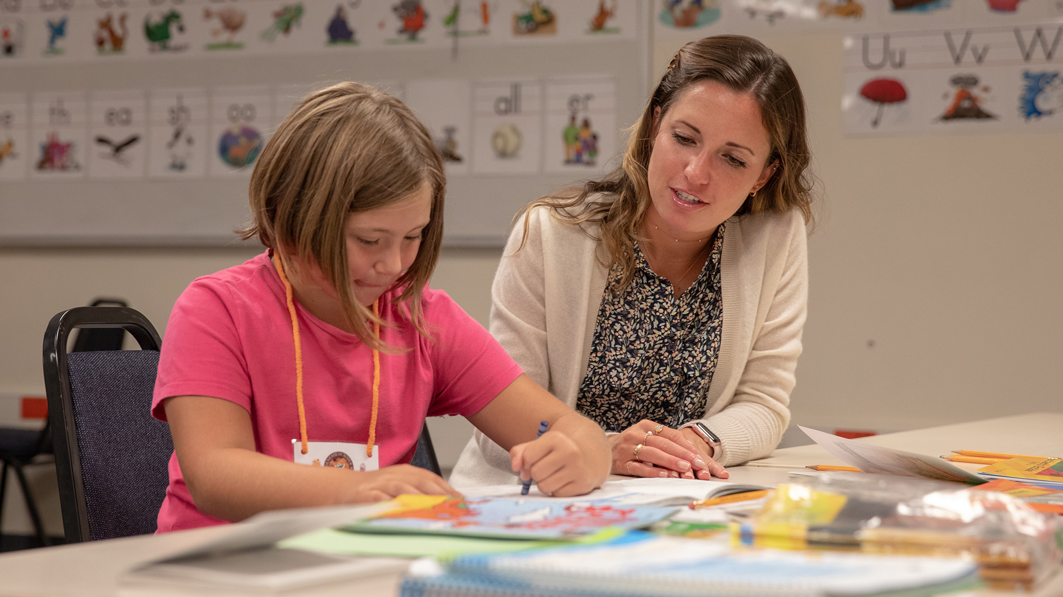 A teacher helps a child color at a classroom table.