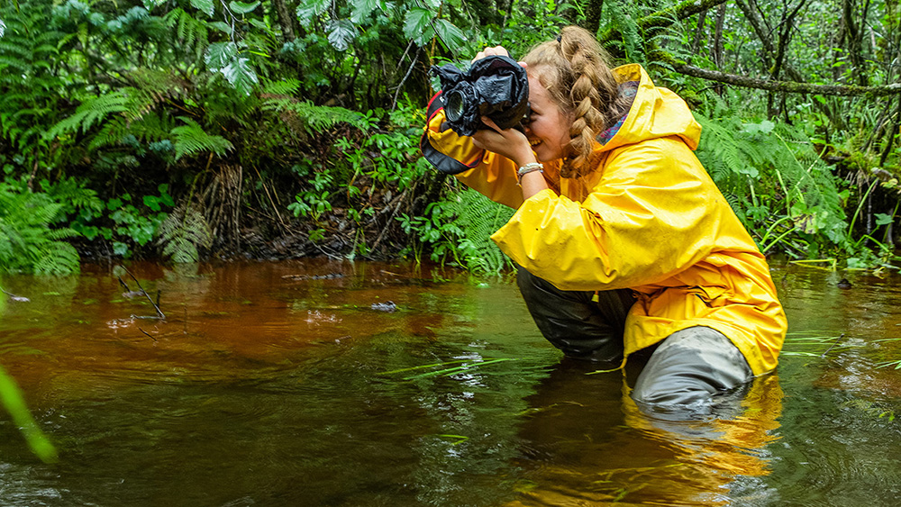 A student standing in a river taking a photo with a camera