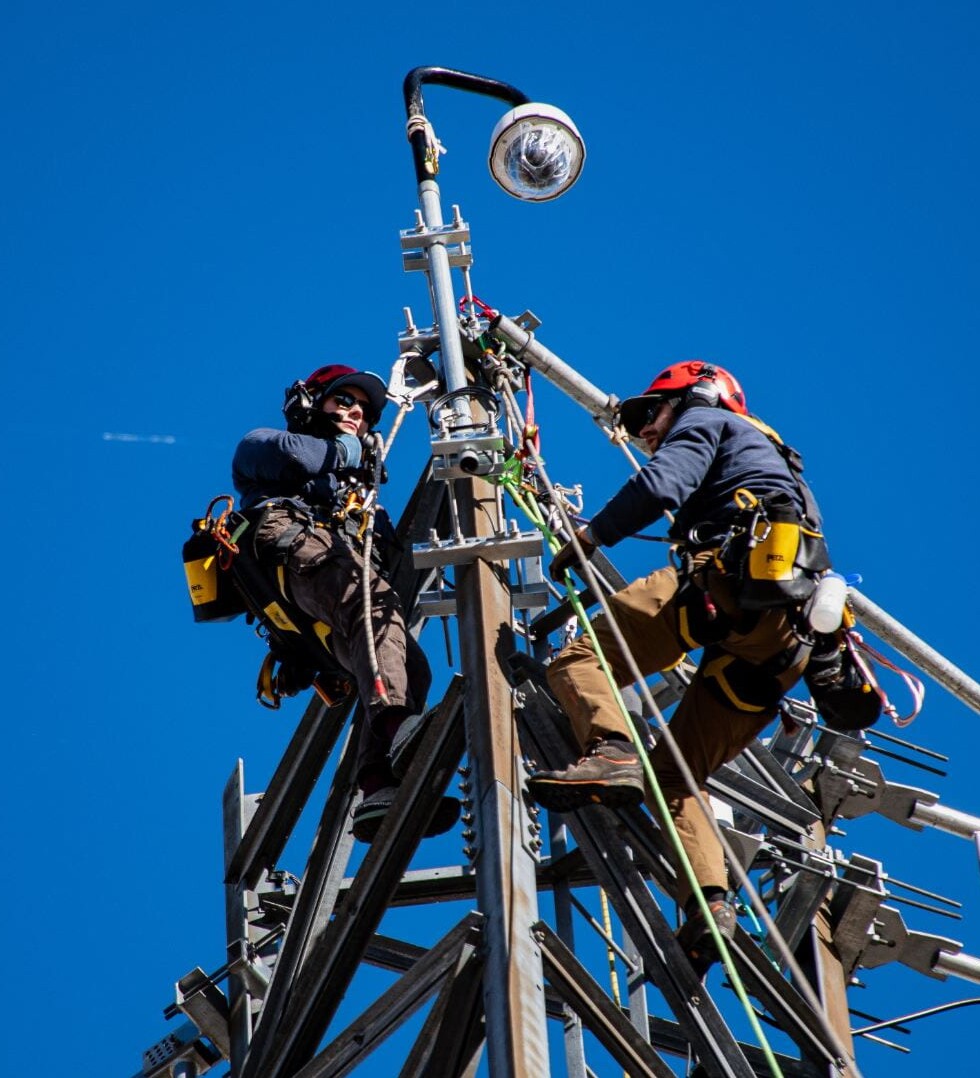 Workers install equipment high on a metal tower beneath a clear blue sky.