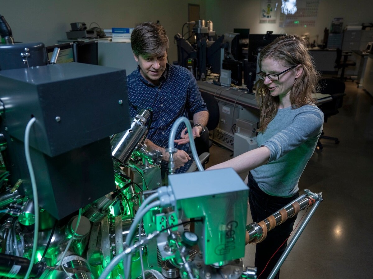 Two researchers operate a green-lit laboratory instrument.