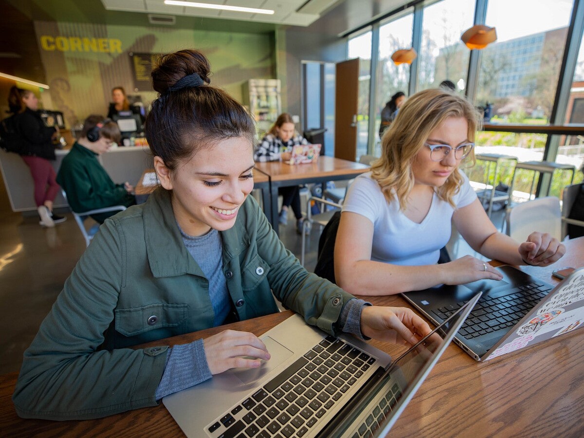 Two students work on laptops at a campus table.