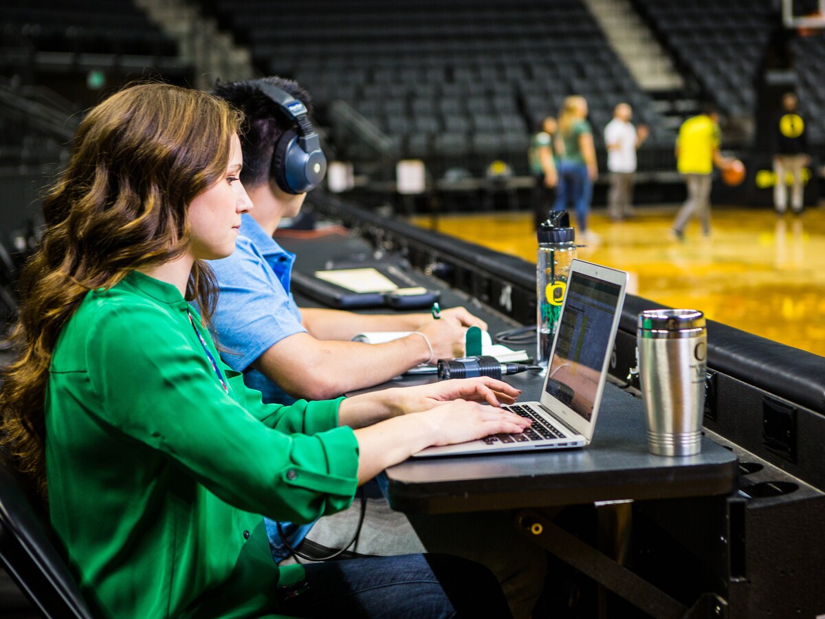 Two students use laptops beside a basketball court.