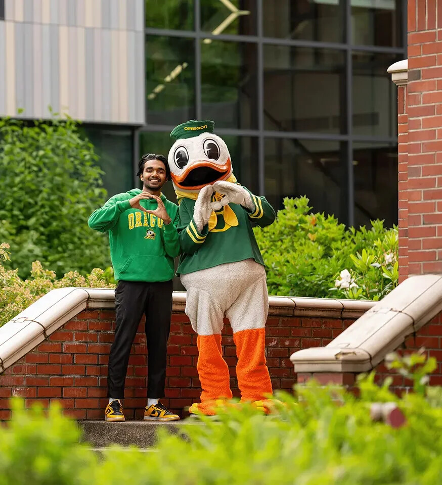 The Oregon Duck and a student make heart shapes with their hands on campus stairs.