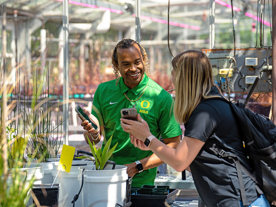 Two students talking and looking at their phones in a greenhouse filled with plants