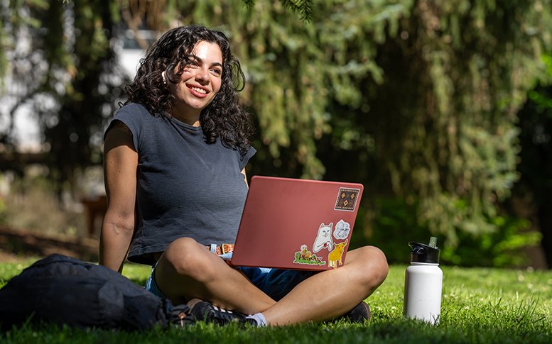 A person smiles with an open laptop while sitting on a campus lawn.