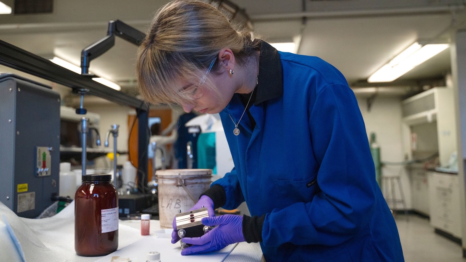 A researcher in a blue lab coat works with an electrochemical cell.