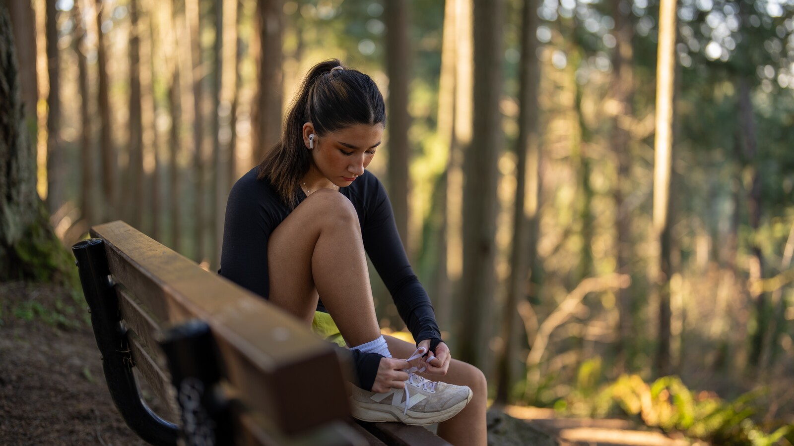 A young woman sits on a forest bench tying a running shoe.