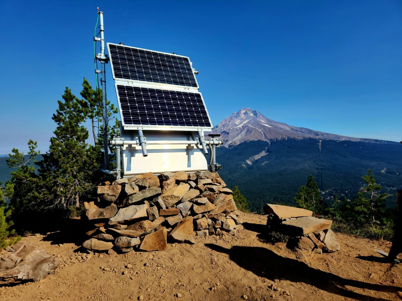 A solar-powered monitoring station stands on a ridgeline with a snow-capped peak in the distance.