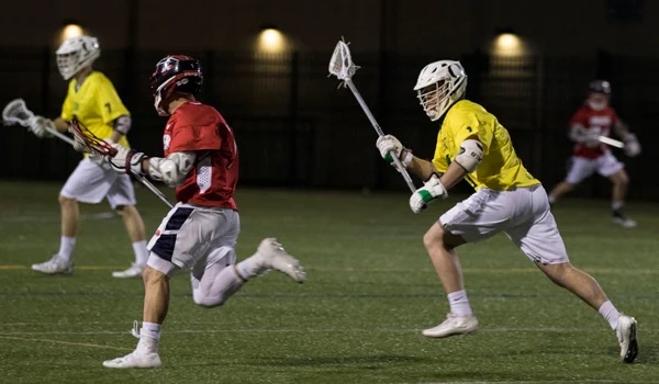Club lacrosse players compete on a turf field at night.
