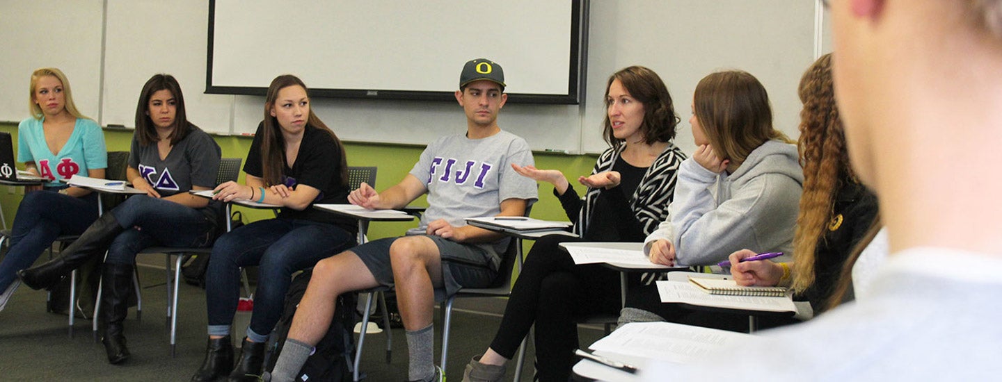 Fraternity and sorority members sit in a classroom meeting, wearing shirts with Greek letters.