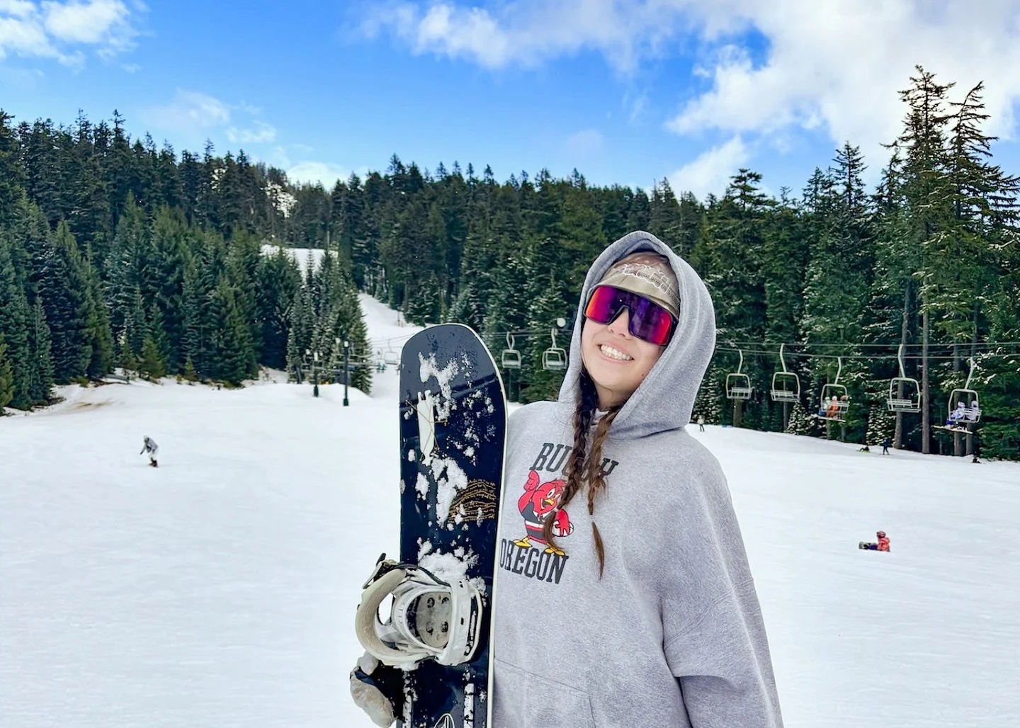 Sydney Seymour stands on a snowy slope holding a snowboard, with a chairlift and evergreens behind her.
