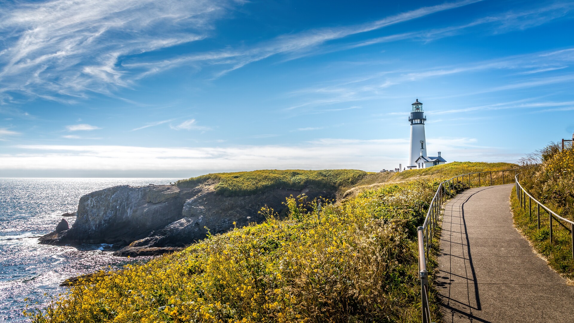 A path curves toward a white lighthouse on a grassy Oregon headland.