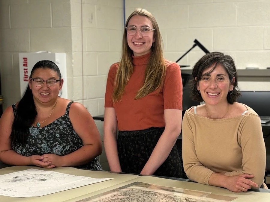 Three people smile behind a table displaying historical maps.